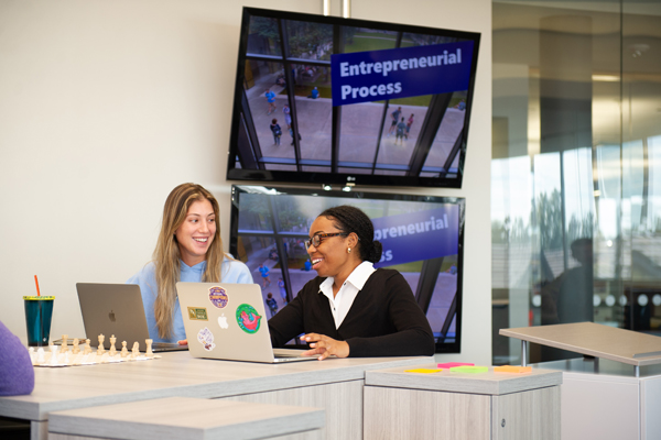 Two women sit behind a table working on a laptop.