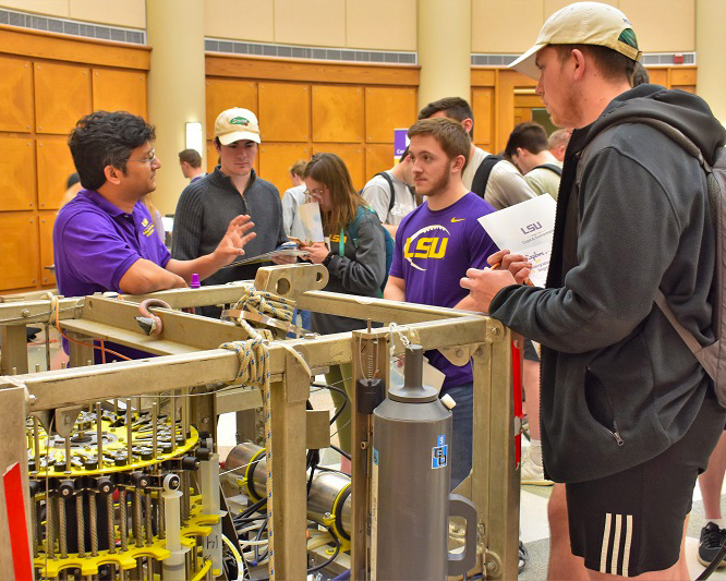 a man stands next to a large metal framed piece of scientific equipment, talking to two students