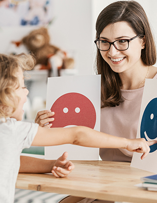 Photo of a young female teacher showing images of a happy face and a sad face to a young child